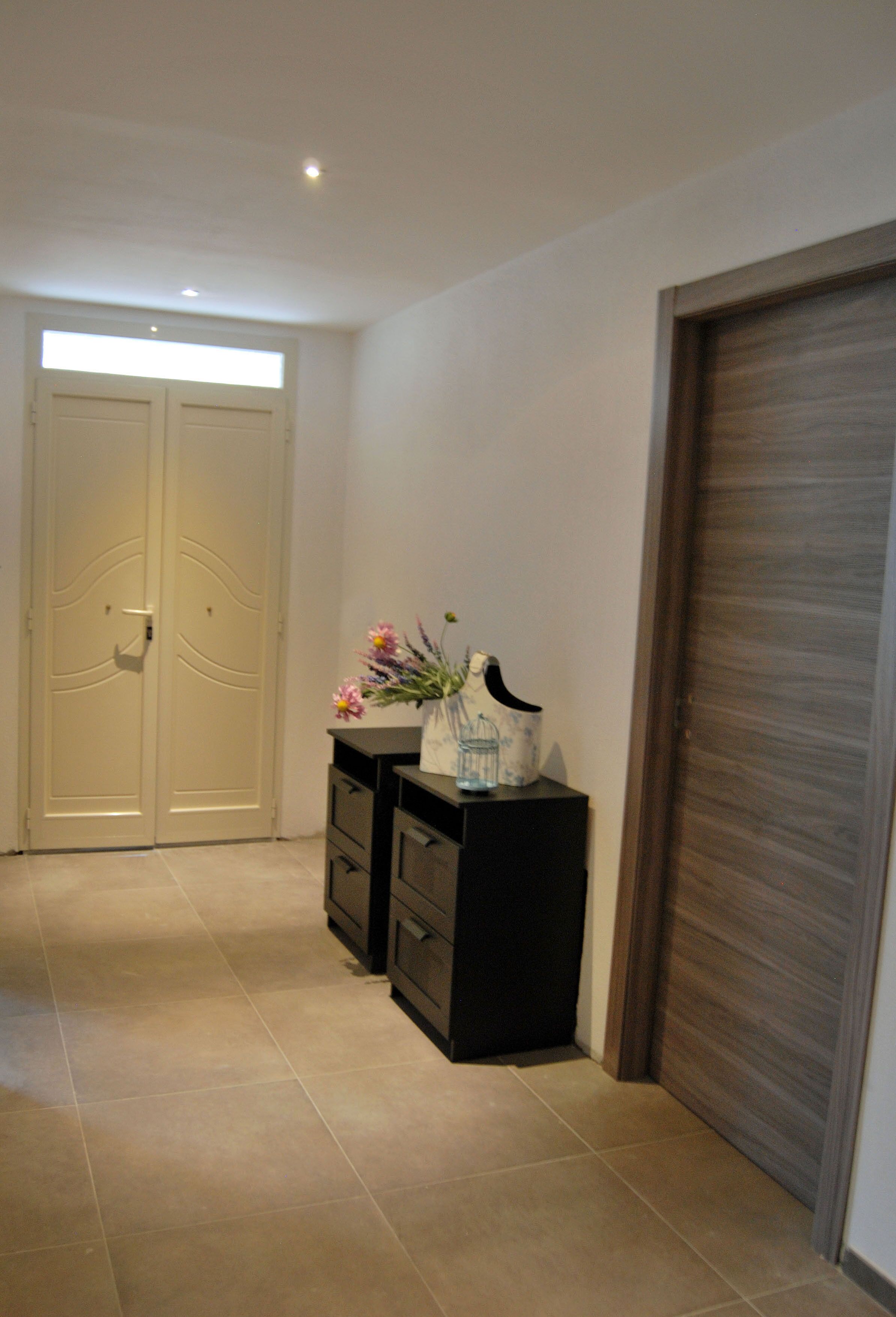 A hallway with beige double doors, a black console table with a vase of flowers, and a wooden door on the right.