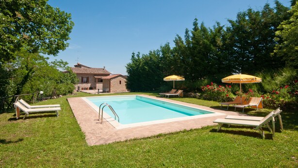 Una piscina con sillones de descanso y sombrillas, rodeada de vegetación y una casa al fondo.
