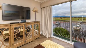 A modern living room with a large flat-screen TV, wooden console, and a view of a parking area and landscape through large windows. - 1 Links Crescent 5 Star Selfcatering House Overlooking Old Course in St Andrews (St Andrews)