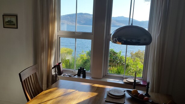A dining area with a table, chairs, a lamp, and a view of the sea through large windows. - Fabulous House with Stunning Sea Views to Skye. By NTS gardens. Great Privacy. (Kyle of Lochalsh)