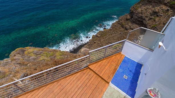 Une terrasse en bois avec une partie recouverte de carreaux bleus, offrant une vue sur une côte rocheuse et une mer d’un bleu limpide.