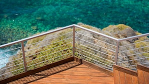 Une terrasse en bois avec des rampes en métal, donnant sur une eau d’un bleu limpide et un paysage rocheux.