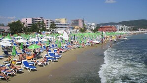 Ein Strand mit Reihen von Liegestühlen und Sonnenschirmen, ein sandiger Uferbereich und Gebäude im Hintergrund.