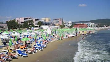 Ein Strand mit Reihen von Liegestühlen und Sonnenschirmen, ein sandiger Uferbereich und Gebäude im Hintergrund.