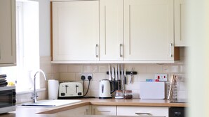 Una cocina con armarios blancos, un tostador, una tetera y un lavabo. Hay un comedor con mantel a cuadros y sillas de madera.