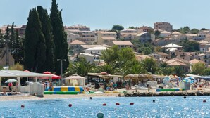 A coastal town with a beach area featuring umbrellas, lounge chairs, and a swimming pool. - Apartment Palm Beach 2 for 6-8 persons - Amazing beach house rental! (Okrug Gornji)