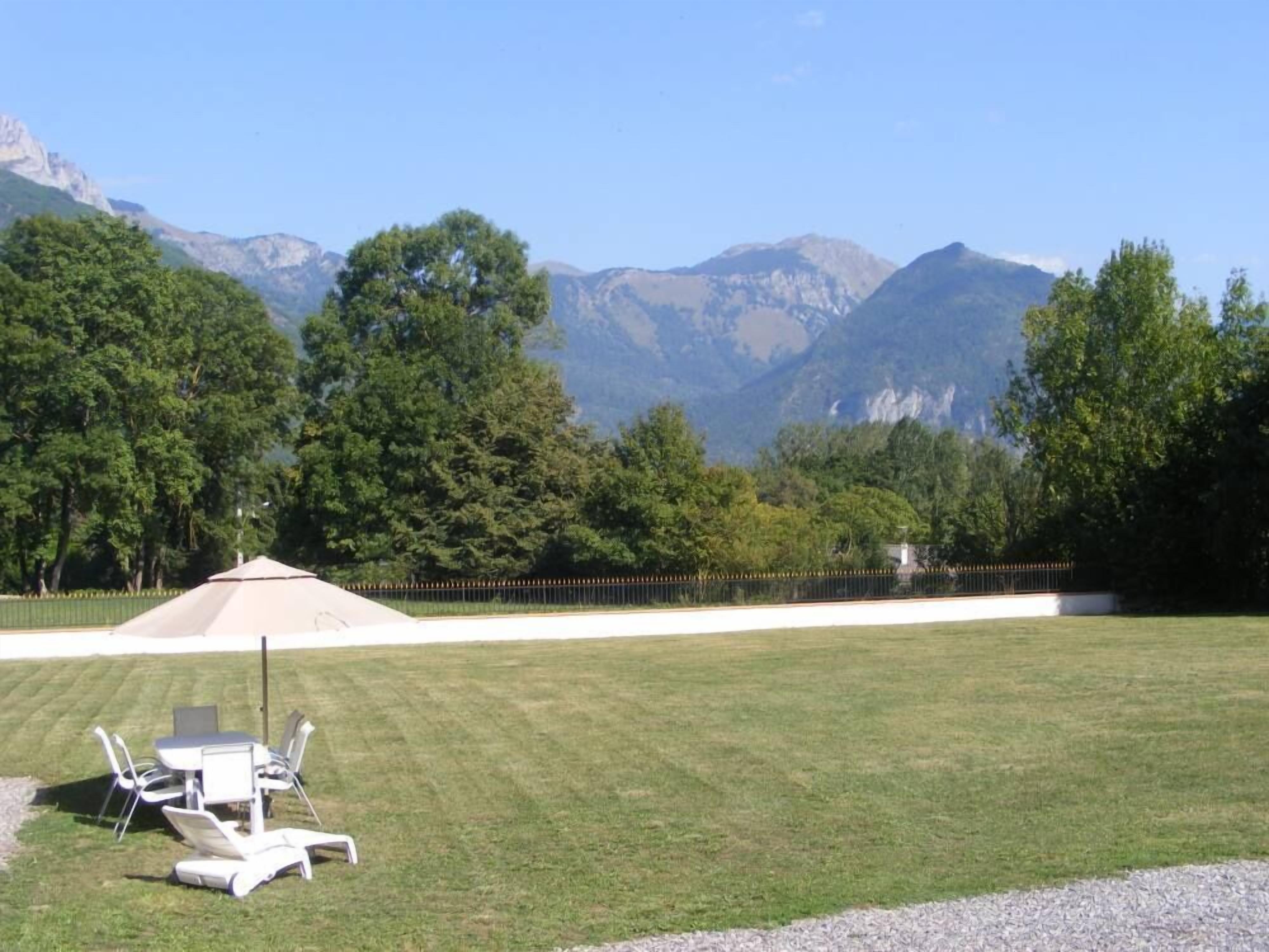 A grassy outdoor area with a dining table and chairs, surrounded by trees and mountains in the background.