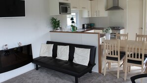 A modern living room with a black leather sofa, a flat-screen TV, and a dining area with wooden chairs and a small table. - MARSHLANDS LAKESIDE NATURE RETREAT (Barton-upon-Humber)