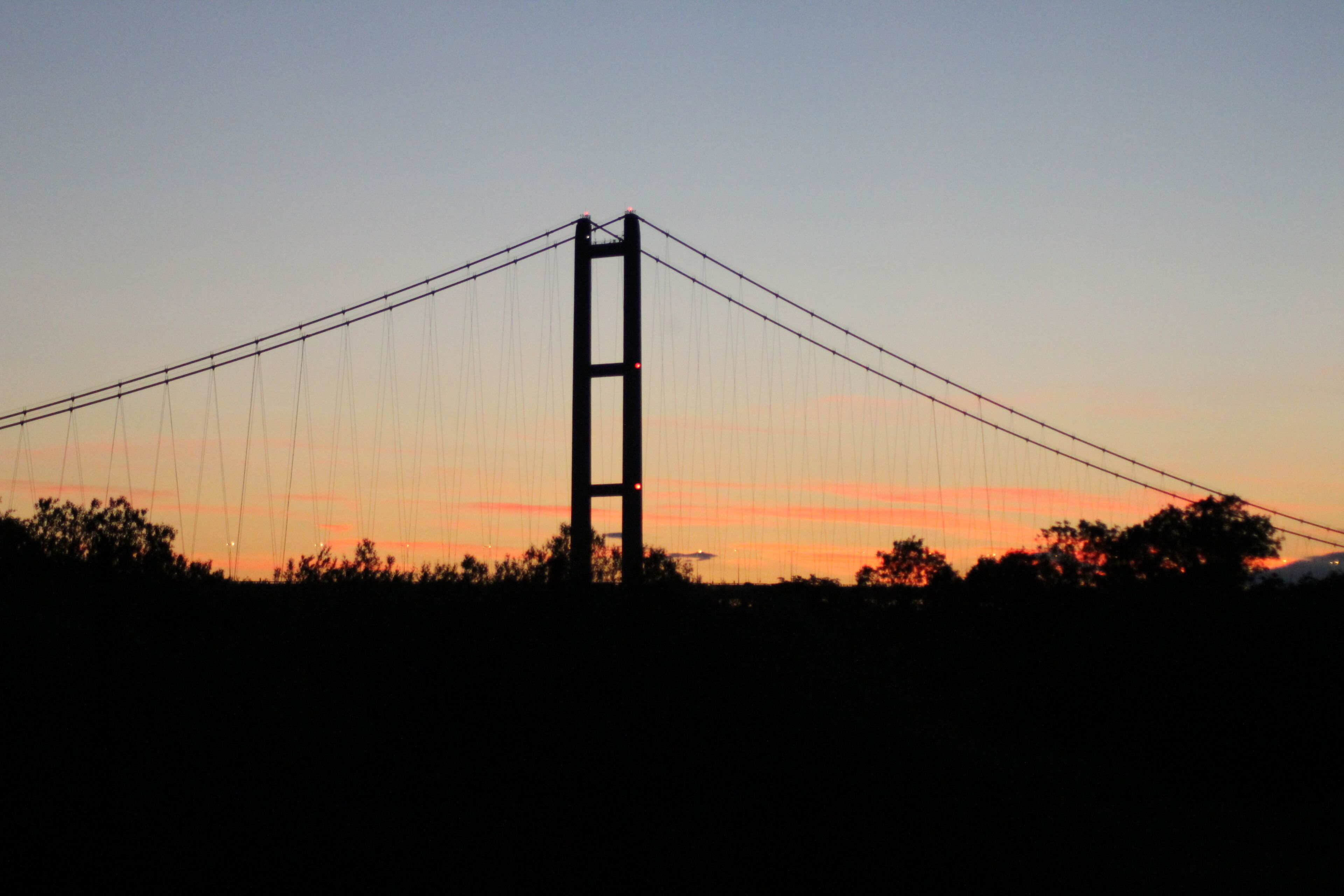 A bridge with a tall central tower and suspension cables against a sunset sky.