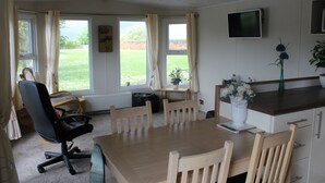 A dining area with a wooden table and chairs, a black office chair, a TV mounted on the wall, and a window with curtains. - MARSHLANDS LAKESIDE NATURE RETREAT (Barton-upon-Humber)