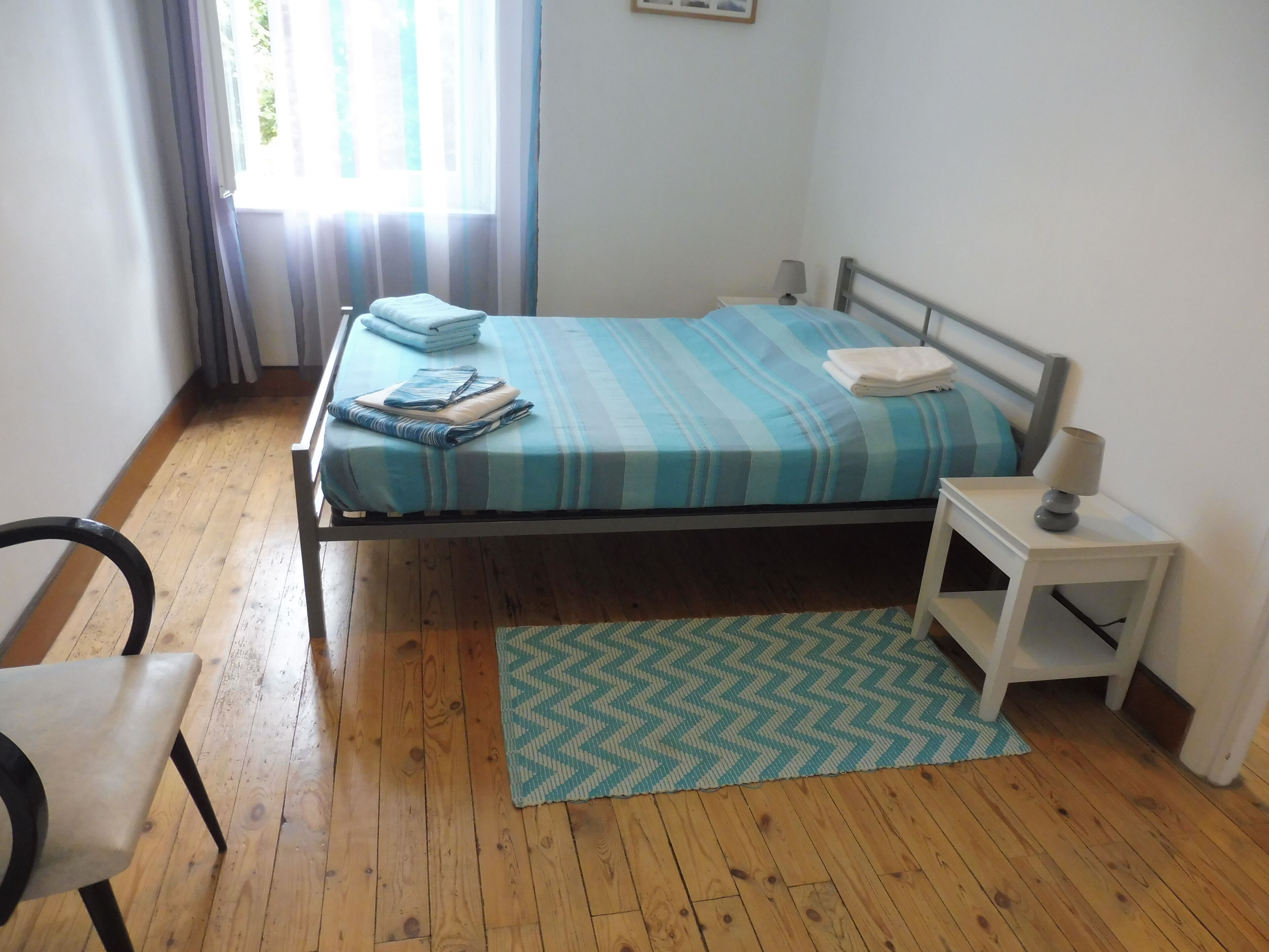 Een slaapkamer met een bed, nachtkastje, stoel en een raam met gordijnen.