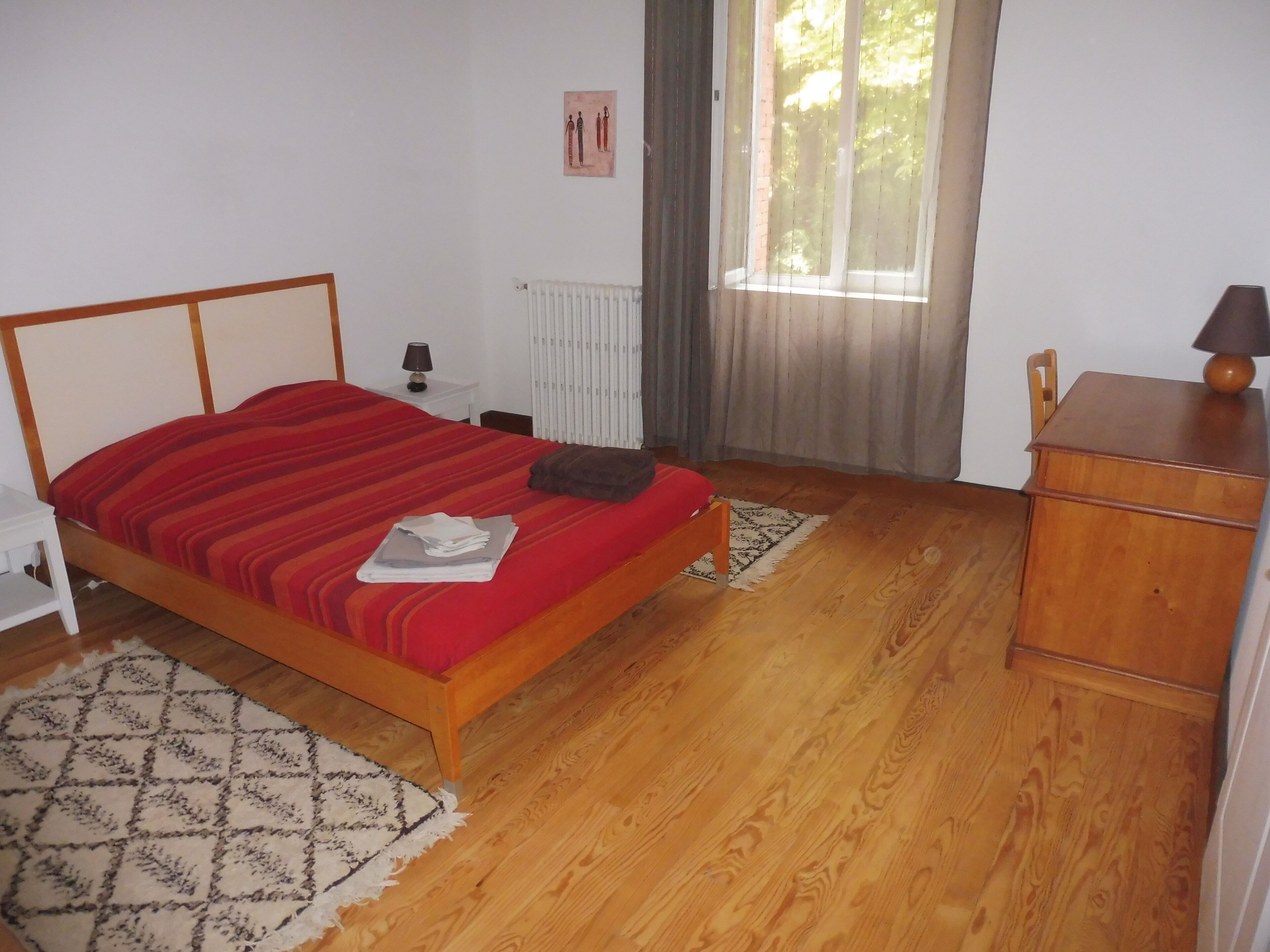 Une chambre à coucher avec un lit en bois, une table de chevet, un radiateur, une fenêtre avec des rideaux et une commode en bois.
