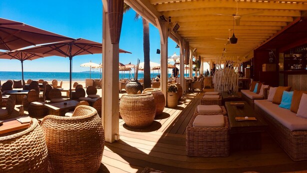 Een terras buiten met rieten meubels en parasols, met uitzicht op het strand.