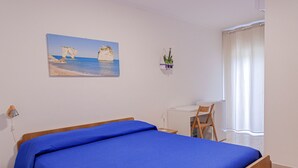 Ein Schlafzimmer mit Bett, Schreibtisch, Stuhl, einem kleinen Tisch, einer Topfpflanze und einem Bild an der Wand.