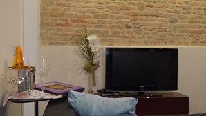 A living room with a stone wall, a flat-screen TV, a small table with a wine glass, a vase with flowers, and a book.