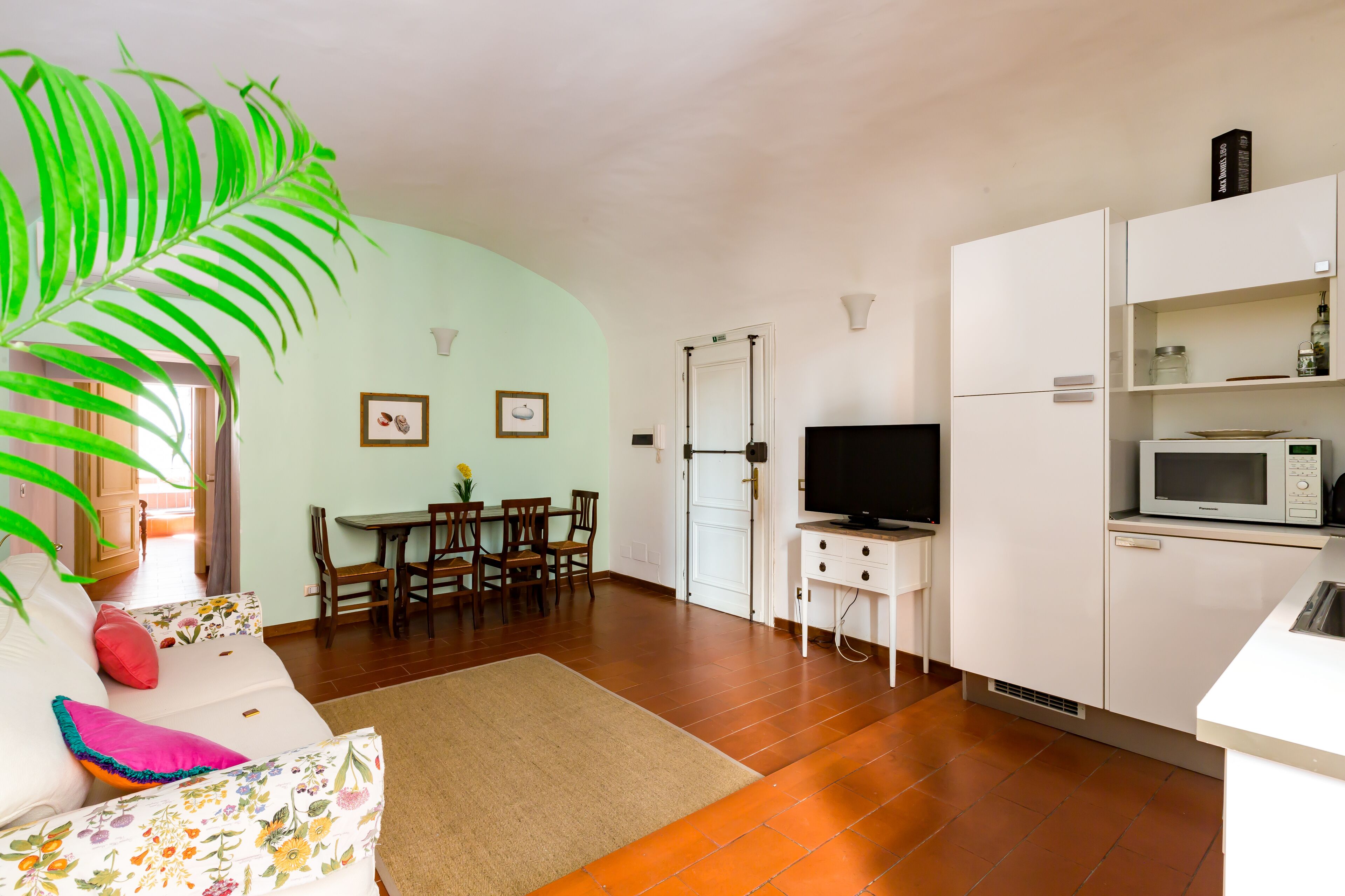 Un salon avec un canapé, une table à manger avec des chaises, un téléviseur, un coin cuisine équipé d’un four à micro-ondes et une grande plante verte.