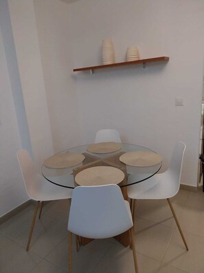 A round glass table with four white chairs, each with a beige cushion. A wooden shelf above holds stacked white plates.