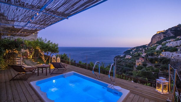 Eine Holzterrasse mit Whirlpool, Liegestühlen und Blick auf das Meer und eine Stadt am Hang.