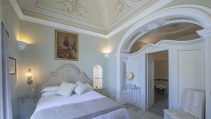 Ein Schlafzimmer mit einem großen Bett, einem Nachttisch, einem Sessel, einem Spiegel und einer Tür zu einem anderen Raum.