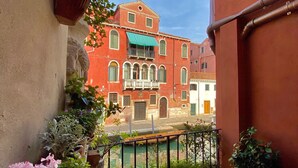 Un balcón con plantas en macetas y vistas a un edificio rojo con ventanas de arco y un toldo verde.