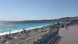 A beach with people sunbathing and swimming, a promenade with blue chairs, and a clear blue sky. - Stunning 2 bed waterfront apartment on Nice Promenade beside the Radisson Hotel (Nice)