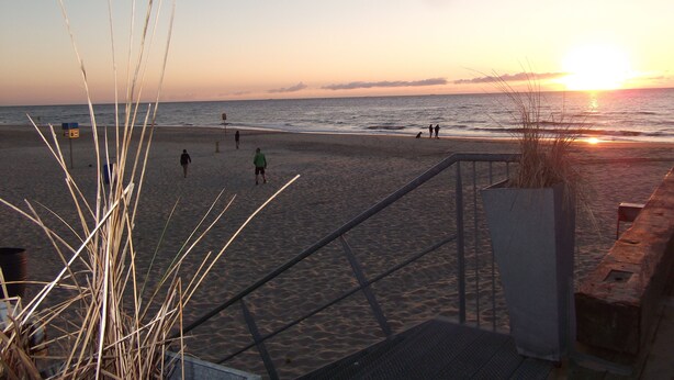 Una playa con un atardecer, gente caminando y una escalera que baja a la playa.