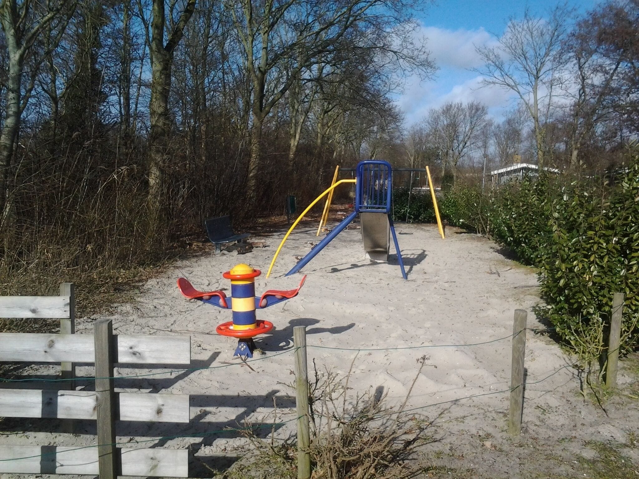 Une aire de jeux avec une balançoire, un toboggan et une structure d’escalade, entourée d’arbres et d’une clôture en bois.