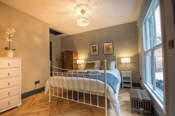 A bedroom with a white metal bed frame, a wooden dresser, a radiator, and a window with blinds.