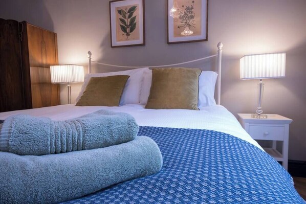 Ein ordentlich bezogenes Bett mit einem blauen Musterüberwurf, zwei Kissen und einer gefalteten Decke. An beiden Seiten des Bettes befinden sich zwei Wandlampen.