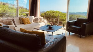 A modern living room with a sofa, armchair, and coffee table, large windows with a view of greenery and hills. - Sun, Sea & Culture. Luxury Villa & Pool. Breathtaking Views to Sea. Inc WiFi (Santa Cristina d'Aro)