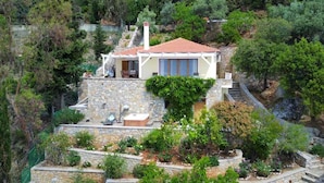 A stone house with a red-tiled roof, a balcony, and a garden with various plants and flowers. - Alonissos/Alonnisos - Idyllic Villa with private beach & jacuzzi,  amazing views (Alonissos)
