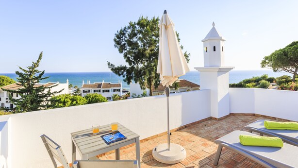 A terrace with a table, chairs, and an umbrella, overlooking a coastal view with buildings and trees. - Sea View !!! 2 bedrooms (Almancil)