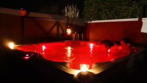 Un jacuzzi con iluminación roja, rodeado por un patio oscuro con un adorno con forma de corazón y una pequeña mesa con una vela.
