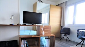 A modern living room with a flat-screen TV on a wooden stand, a bookshelf with books, a blue sofa, a coffee table with decorative items, and a view of the outside through a window. - Studio 3*, SEA VIEW and facing the dragon, 150m from the BEACH, near the 2 caps site (Calais)