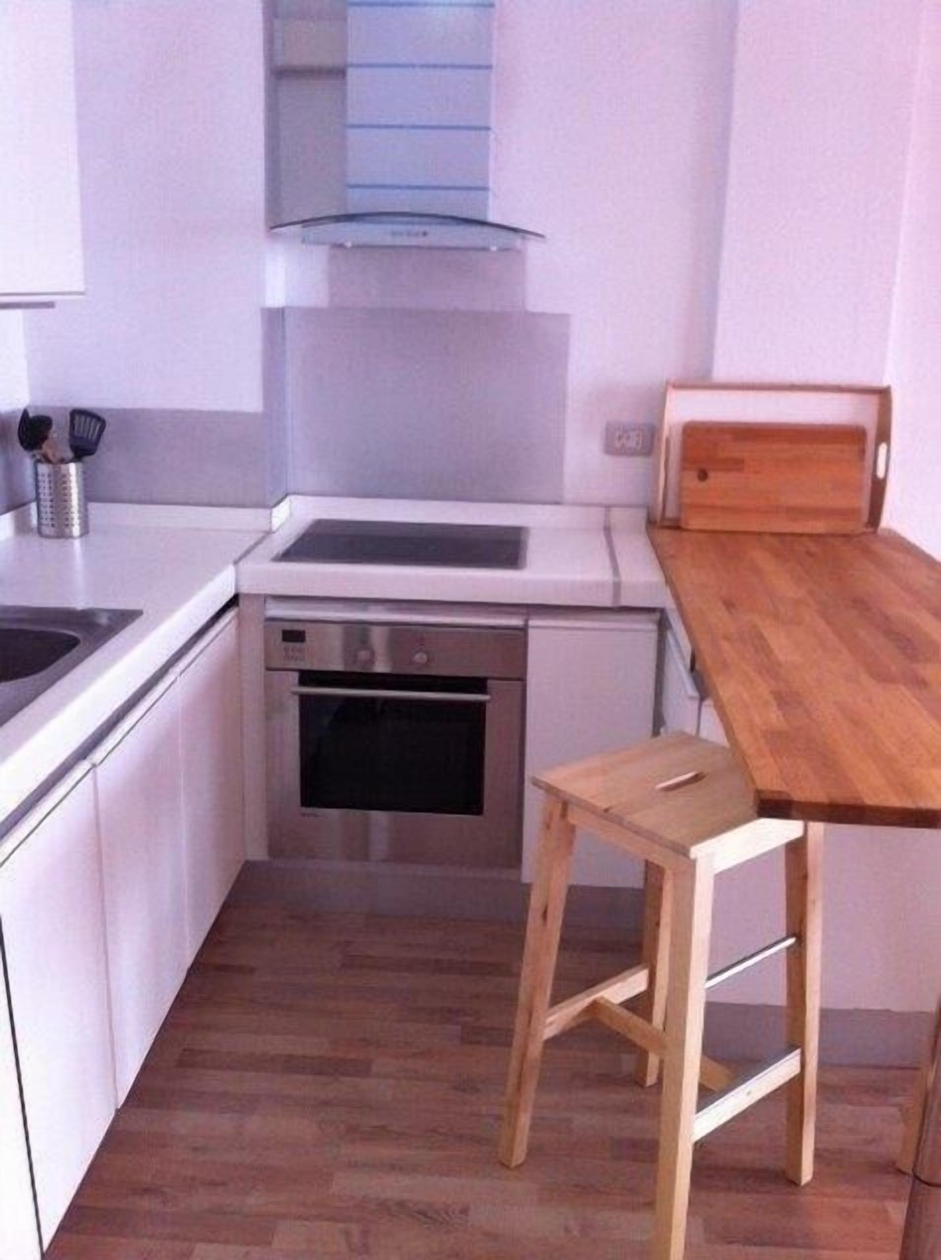 Una cucina compatta con pensili bianchi, forno integrato, uno sgabello di legno e un piccolo tavolo in legno.