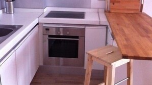 Cocina compacta con armarios blancos, horno empotrado, un taburete de madera y una pequeña mesa de madera.