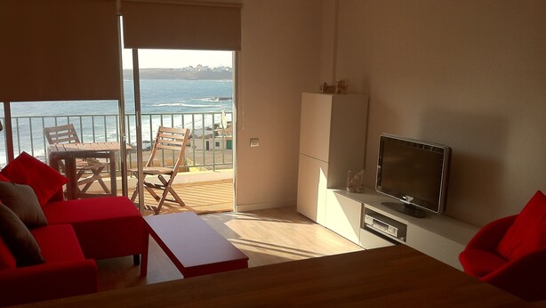 Una sala moderna con una mesa de madera, un sofá rojo y un televisor sobre un mueble bajo. Se ve un balcón con vistas al mar y edificios.