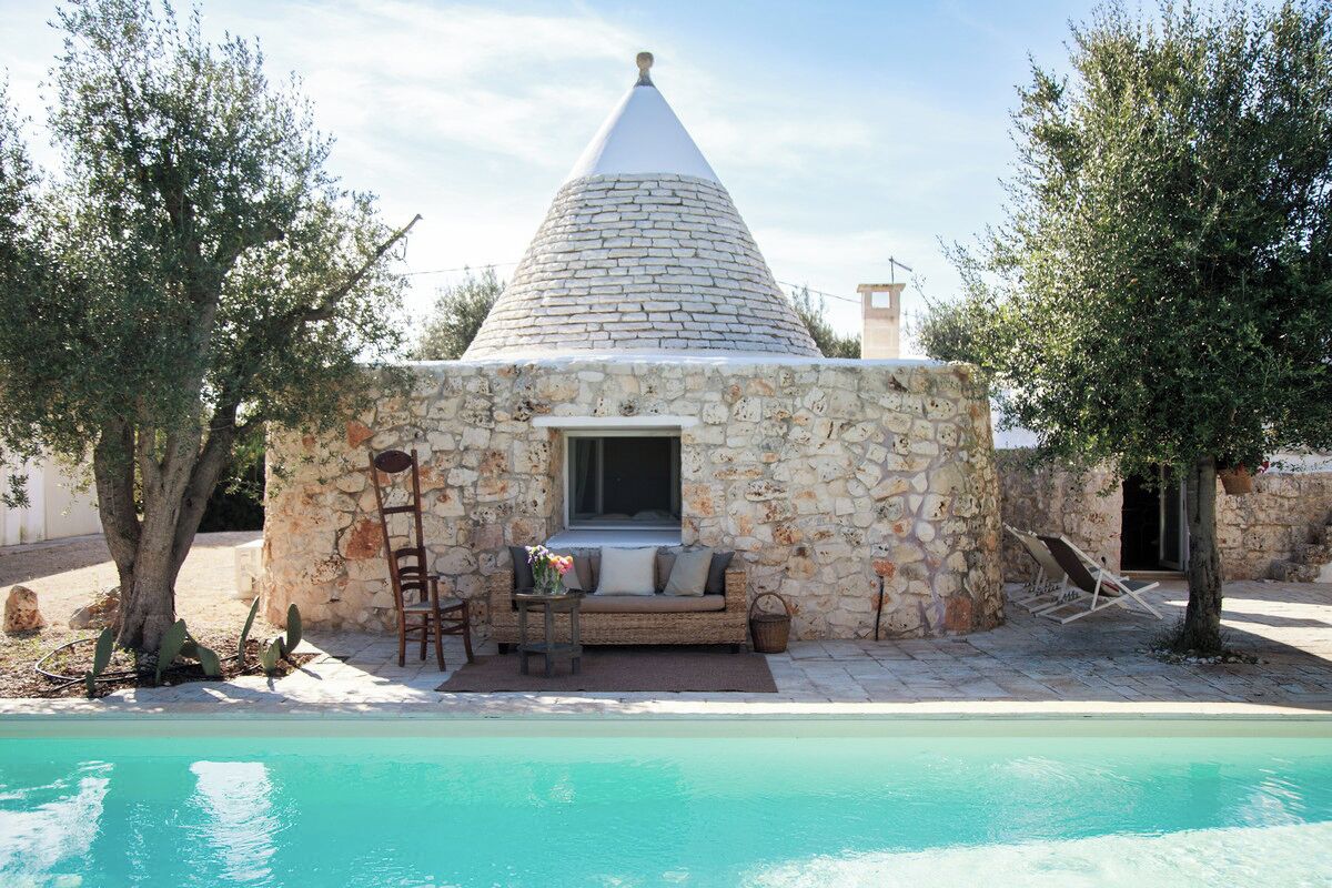 Une maison en pierre avec un toit conique, un petit porche doté d’un canapé et de chaises, et une piscine en premier plan.