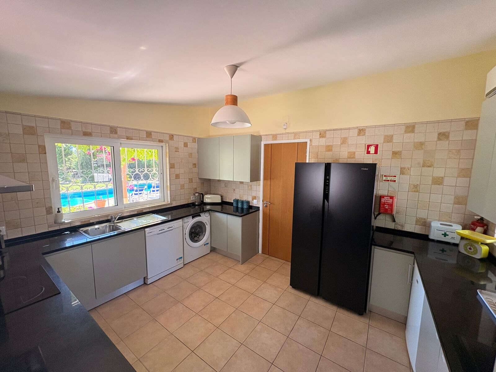 Een kleine keuken met een wasmachine, een koelkast en een raam met uitzicht op groen.