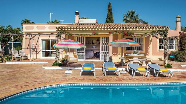Zona piscina con lettini e ombrelloni, una piscina e un edificio con tetto in tegole e camino.