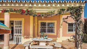 Un patio con tavolo apparecchiato per mangiare, una zona coperta con una pergola e un edificio con grate alle finestre.