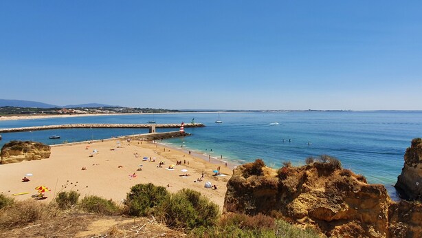 Una playa con aguas azules cristalinas, afloramientos rocosos y un muelle con barcos.