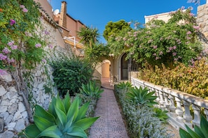 A narrow pathway flanked by lush vegetation and stone walls, leading to a building entrance.