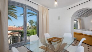 A modern room with a glass table, white chairs, a bed, and a view of palm trees and buildings.