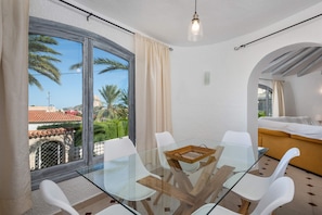 A modern room with a glass table, white chairs, a bed, and a view of palm trees and buildings.