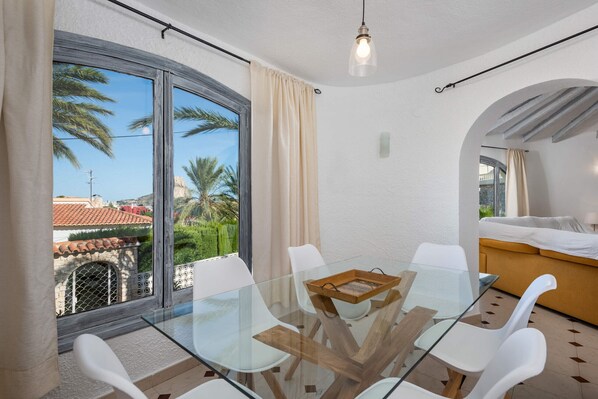 A modern room with a glass table, white chairs, a bed, and a view of palm trees and buildings.