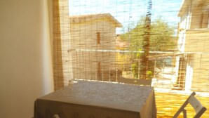 Un balcón con una mesa y dos sillas, una ventana con mosquitero y vistas a edificios y árboles.