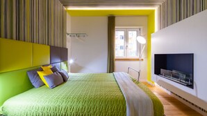 A modern bedroom with a green bedspread, a flat-screen TV mounted on the wall, and a window with curtains.