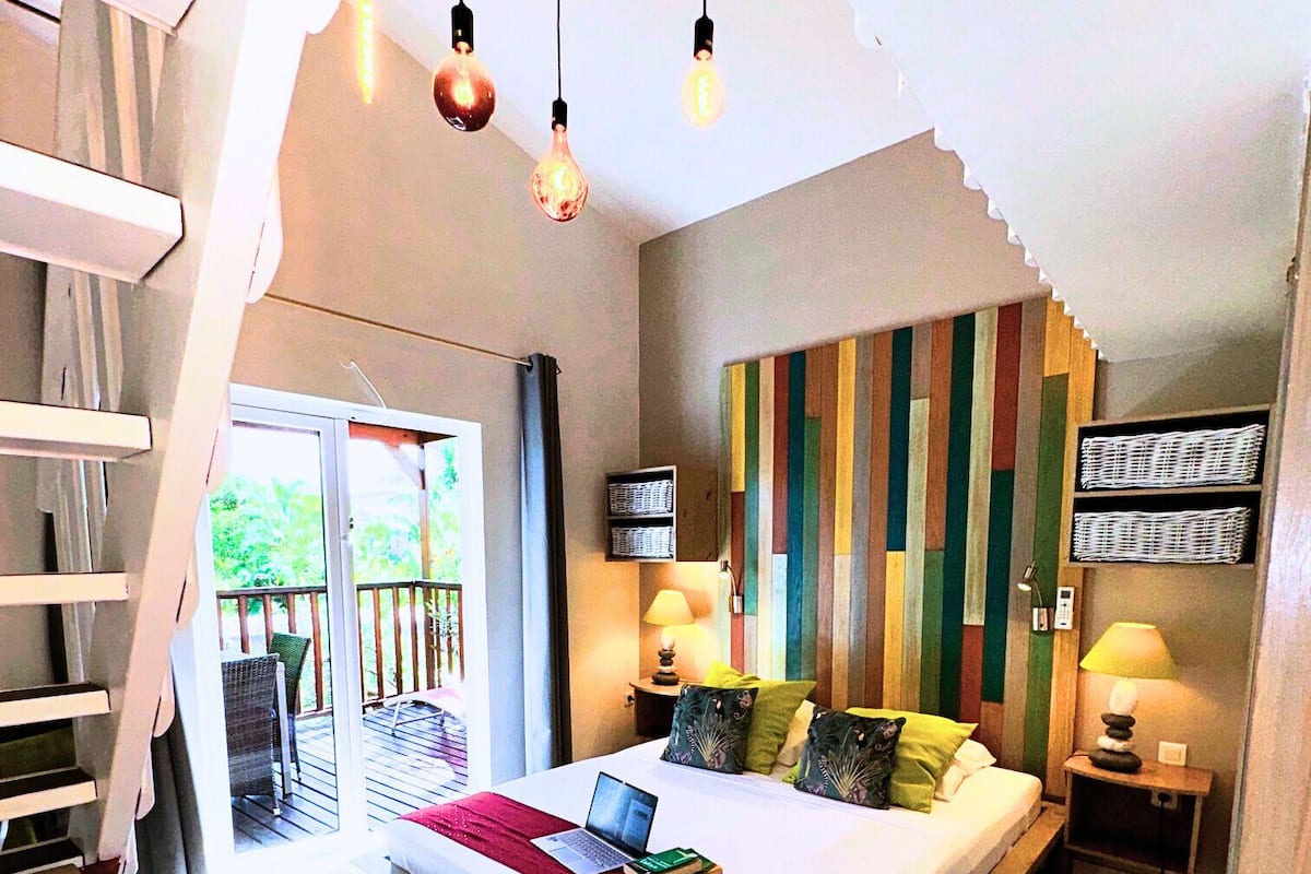 A bedroom with a bed, a laptop, a bedside table, a lamp, and a view of the outdoors.