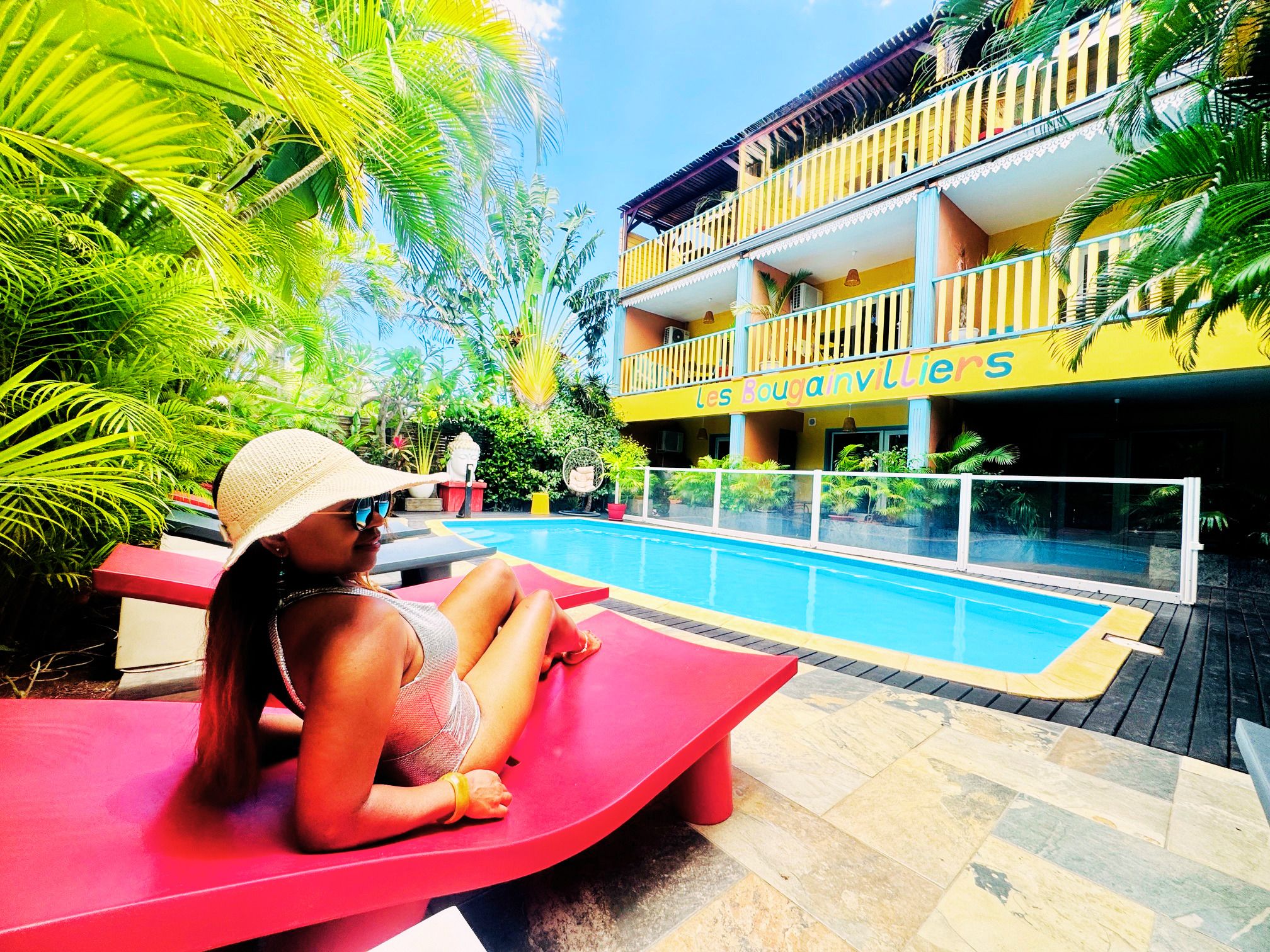 A woman sitting on a red lounge chair by a poolside, with a building labeled 'Les Bougainvillers' in the background.